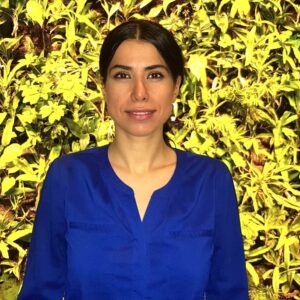 Farinaz Basmechi stand in front of a green leafy wall in a blue shirt facing the camera
